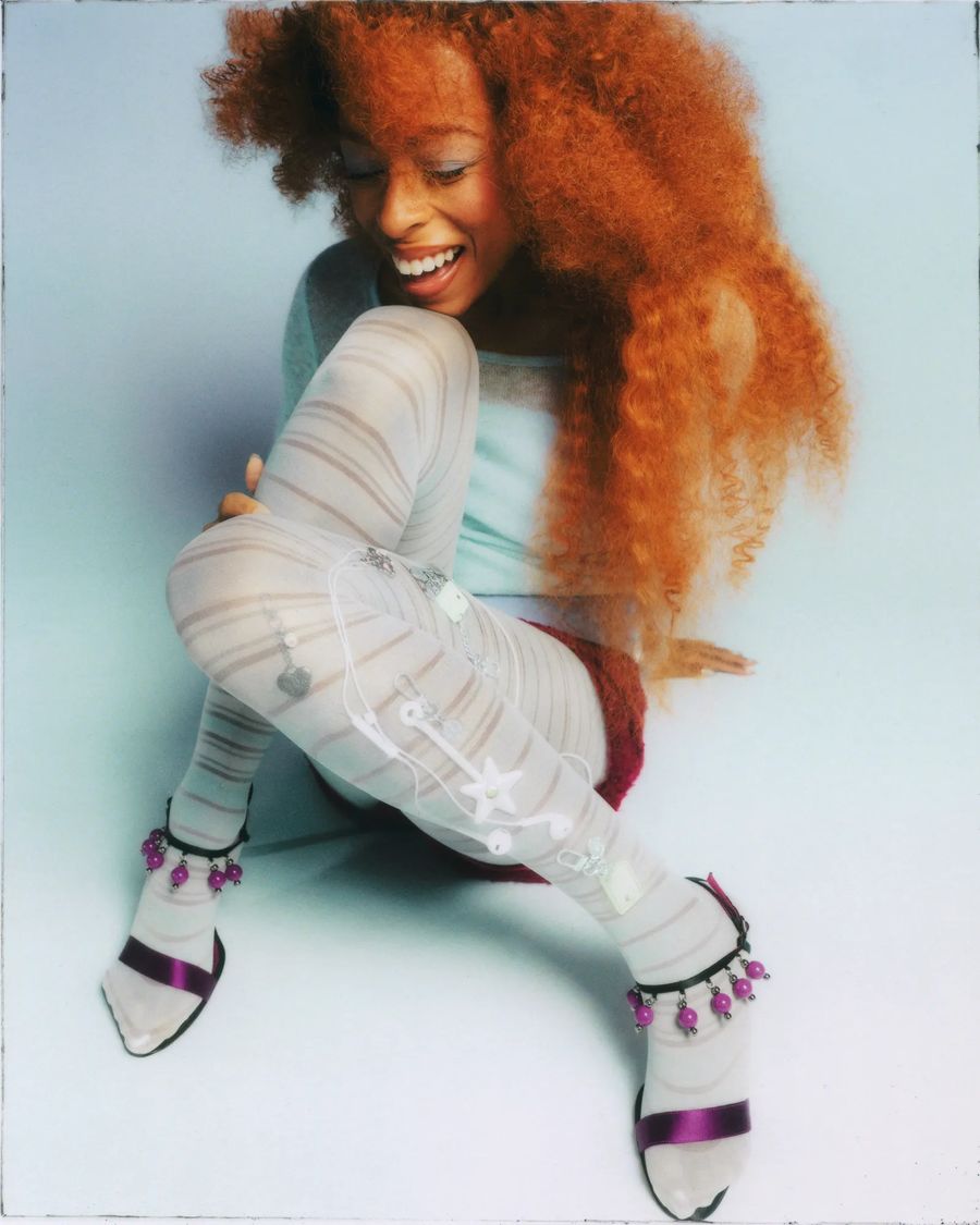 Ravyn Lenae laughs, a Wand and Mini Cards clipped to her striped tights